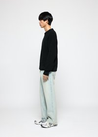 Ung mann i svart genser, lyseblå jeans og hvite joggesko står i profil mot en lys hvit bakgrunn.