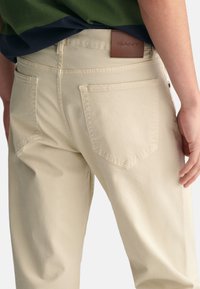 Beige bomullsbukser med baklommer, en brun lærlabel fra GANT på midjen, og en avslappet passform.