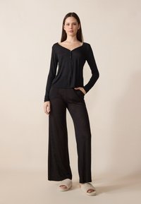 Μαύρη μπλούζα με μακριά μανίκια, V λαιμόκοψη και κουμπί, συνδυασμένη με μαύρο παντελόνι φαρδιάς γραμμής και ανοιχτόχρωμες αφράτες παντόφλες. Λείος υφασμάτινος καμβάς.