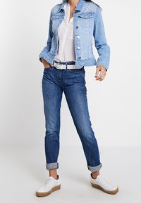 Ljusblå denimjacka med nitar, vit blus, mörkblå jeans och vita sneakers med en tan-färgad sula. Krage och knäppningar.