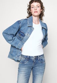 Lys blå denimjakke med klassisk snitt, med to brystlommer, knappestengning og litt slitt tekstur; kombinert med en hvit topp.