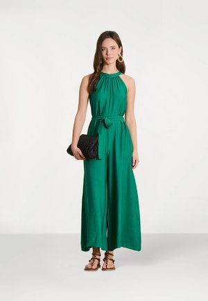Femme portant une combinaison verte sans manches à col halter avec un lien à la taille, tenant une pochette noire, et portant des sandales marron à lanières ainsi que des boucles d'oreilles dorées.