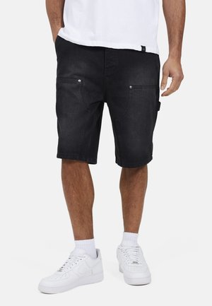 Uomo che indossa pantaloncini di jeans neri, calzini bianchi e scarpe da ginnastica bianche, in piedi davanti a uno sfondo semplice.