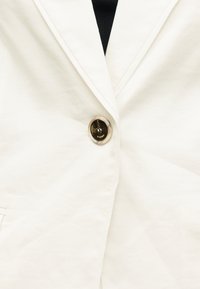 Λευκό σακάκι με ένα μόνο σκούρο, στρογγυλό κουμπί κλειστό στο κέντρο πάνω από ένα μαύρο τοπ.