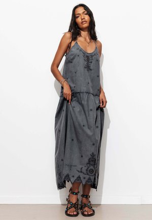 Femme debout contre un mur blanc portant une robe longue ample gris foncé avec une broderie florale et des sandales noires avec des touches de bleu.