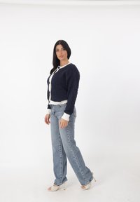 Cardigan lavorato a maglia blu navy con rifiniture e bottoni bianchi, abbinato a jeans azzurri a vita alta e sandali beige con tacco.