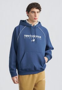Jonge man die een blauwe New Balance Athletics hoodie en mosterdkleurige broek draagt, staand met één hand in zijn zak tegen een effen achtergrond.