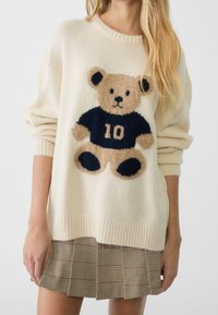 Kvinna iförd en krämfärgad tröja med en stor teddybjörn som bär en marinblå skjorta med nummer 10, tillsammans med en plisserad rutigt kjol.