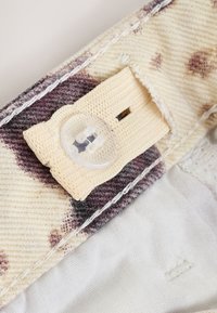 Tissu en coton avec une base beige clair et des motifs tachetés marron. Comprend une fermeture à bouton cousu et une sangle texturée en forme triangulaire.