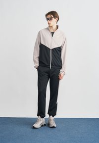 Αθλητική φόρμα Nike με ανοιχτό γκρι και μαύρο σχέδιο, διαθέτει μπουφάν με φερμουάρ και παντελόνι με ελαστική μέση, συνοδευόμενη από γκρι αθλητικά παπούτσια.