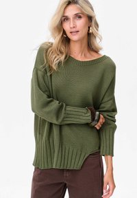 Przesadnie duży zielony sweter z dzianiny z prążkowanym dekoltem i mankietami, z wysokim i niskim dołem oraz teksturowanym materiałem. Połączony z brązowymi spodniami.