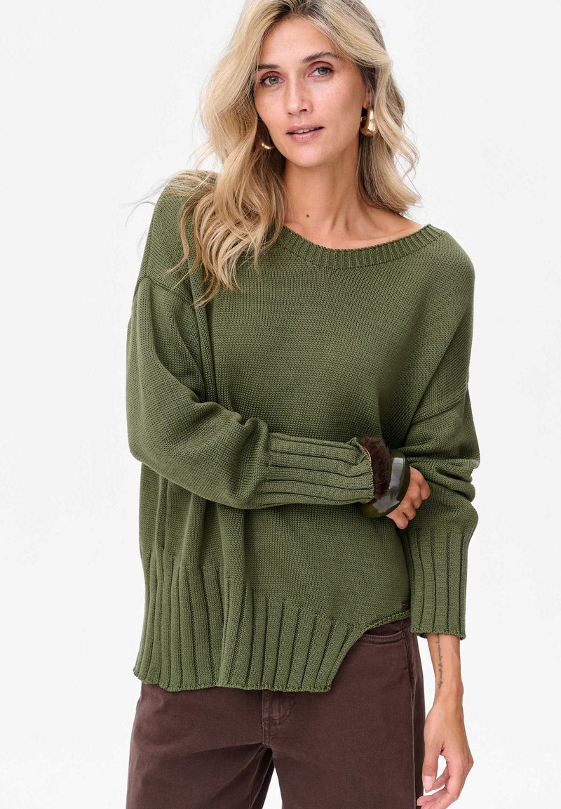 Przesadnie duży zielony sweter z dzianiny z prążkowanym dekoltem i mankietami, z wysokim i niskim dołem oraz teksturowanym materiałem. Połączony z brązowymi spodniami.