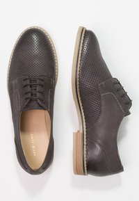 Scarpe in pelle marrone scuro perforata con punta arrotondata, design con lacci e suola di un marrone chiaro a contrasto. Texture liscia con cuciture minime.