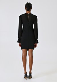 Robe noire avec de longues manches transparentes, une silhouette ajustée et un dos en keyhole. Fini avec une fermeture éclair et une texture douce. Portée avec des talons noirs.