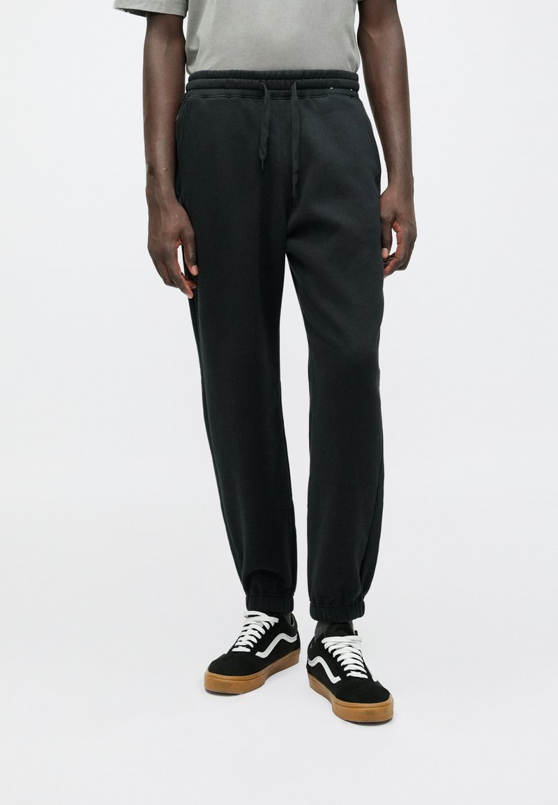 Czarne dresy z elastycznym pasem i mankietami, o gładkiej fakturze. Noszone z czarnymi i brązowymi sneakersami.