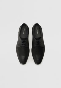 Chaussures noires en cuir avec une texture lisse et un motif nervuré, dotées d'un bout pointu et de lacets noirs. Logo visible sur la semelle intérieure.