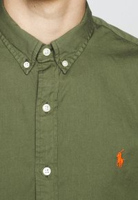 Olivgrön skjorta med knäppning i bomullstyg. Har en klassisk krage, vita knappar och en orange broderad logotyp på bröstet.