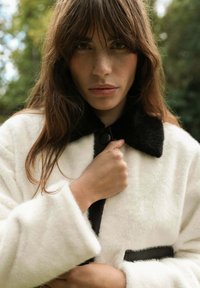 Manteau en fourrure blanche avec un col noir, doté d'une fermeture à boutons et d'une texture lisse ; le modèle tient le manteau autour de la poitrine.