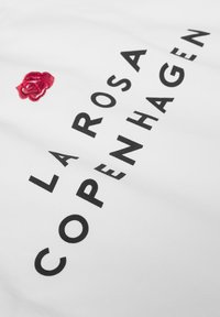 Textil blanco con una rosa bordada en rojo y un texto en negrita negro que dice "LA ROSA COPENHAGUE". Textura suave, diseño minimalista.