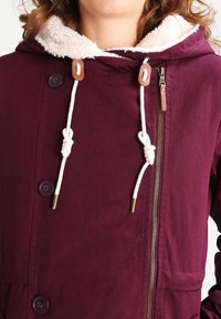 Burgunderfarvet lang jakke med blød cremefarvet hættemaling, med en tovejslynlås, snore og brune læderdetaljer.