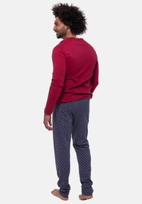 Rød langærmet skjorte parret med marineblå mønstrede pyjamasbukser med lyserøde og hvide prikker. Modellen står barfodet og viser en afslappet pasform.