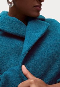 Abrigo de lana turquesa con un gran cuello, textura gruesa y diseño cruzado. Se ve una mano sujetando la tela.