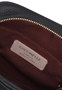 Indvendig på en sort lædertaske med en burgunder stofforing. Har et lille mærke med teksten "Coccinelle Autentisk Italiensk Læder." Guld lynlås.