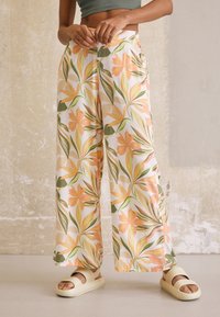 Pantalon à jambes larges en tissu léger avec un motif floral mettant en avant des tons pêche et verts, associé à des sandales slides crème.