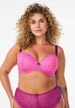 Donna con capelli ricci che indossa un reggiseno in pizzo rosa e mutandine in pizzo viola, in piedi con una mano sul fianco, sfondo neutro.