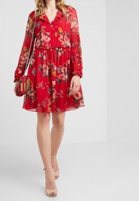 Femme portant une robe rouge à fleurs, longueur genou, à manches longues, tenant un sac à main rouge et des escarpins nus à brides de cheville dans un studio blanc.