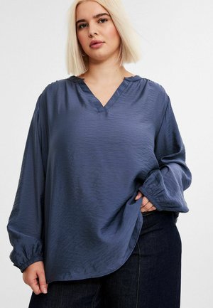 Blå långärmad blus med V-hals, tillverkad av lätt, texturerad tyg. Har elastiska ärmslut och en avslappnad passform. Bärs med mörka jeans.