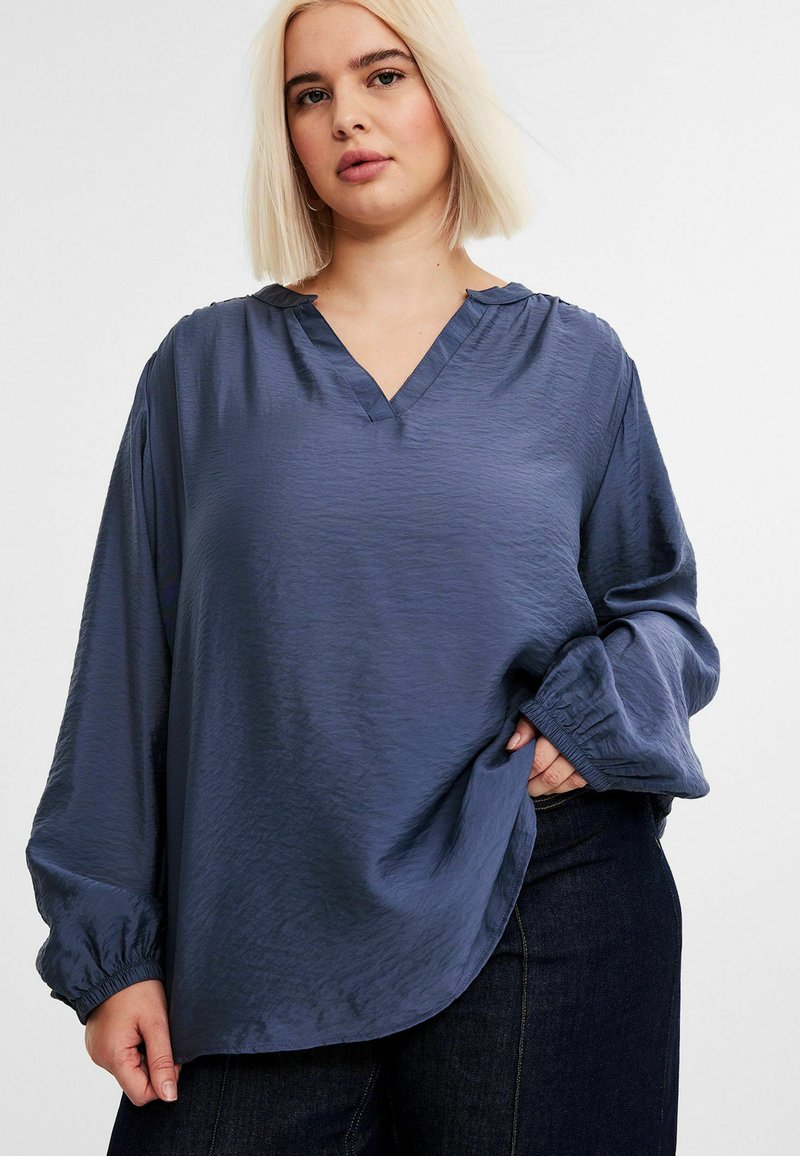 Blå, langærmet bluse med V-hals, lavet af letvægts tekstureret stof. Har elastiske manchetter og en afslappet pasform. Båret med mørke jeans.