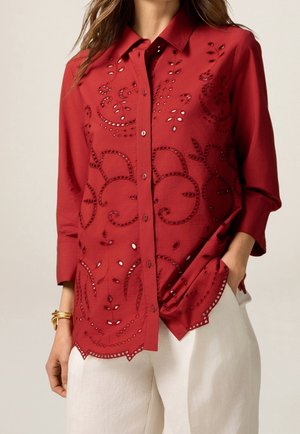 Femme portant un chemisier rouge à boutons avec un motif floral brodé découpé, associé à un pantalon blanc et des bracelets dorés au poignet gauche.