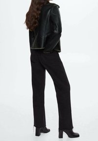 Vrouw in een zwart leren jack, zwarte high-waist jeans met rafelige mannen en zwarte heeled enkelboots op een witte achtergrond.