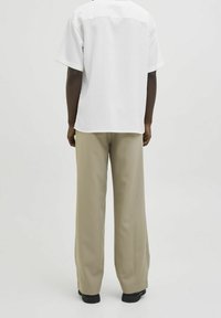 Uomo che indossa pantaloni beige a gamba larga, scarpe nere e una camicia bianca a maniche corte con sottili righe verticali, visto da dietro.