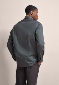 Schwarzes Hemd mit weißen Punkten und metallischem Glanz, mit einem braunen Kragen, langen Ärmeln und abgerundetem Saum. Getragen mit grauen Hosen.