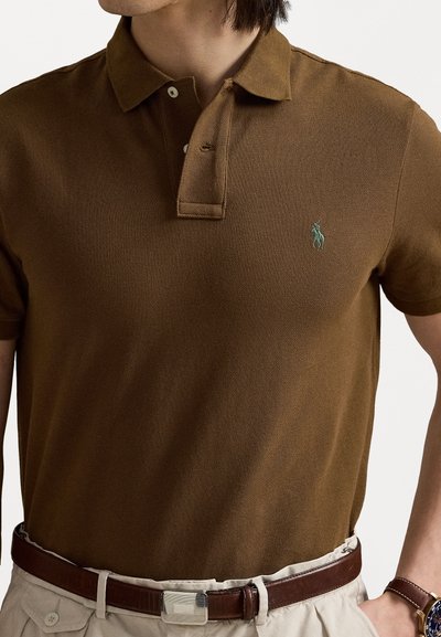 Tricou polo maro cu un model texturat, guler cu două butoane și un mic logo verde; purtat cu o curea maro și pantaloni de culoare deschisă.