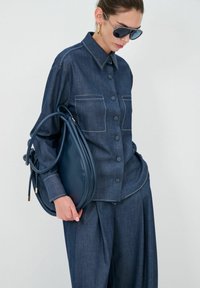 Denim overhemd met een knoopsluiting aan de voorkant, twee borstzakken en contrasterende witte stiksels. Houdt een marineblauwe leren tas vast met een afgeronde vorm en dubbele handvatten.