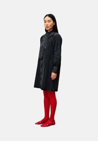 Vrouw die staat in een zwarte denim jurk met lange mouwen en een stropdas, rode panty’s en rode platte schoenen, tegen een witte achtergrond.