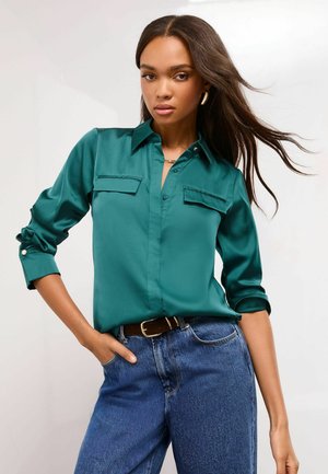 Türkisfarbenes Satinhemd mit langen Ärmeln, zwei Brusttaschen und aufgerollten Manschetten, kombiniert mit hochsitztenden blauen Jeans.