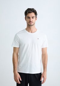 T-shirt bianco in cotone con colletto rotondo e maniche corte, caratterizzato da un piccolo logo colorato sul petto. Indossato con pantaloni neri.
