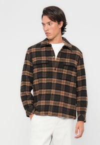 Brown and black plaid flannel shirt with a collared neckline, two chest pockets, and a relaxed fit, paired with light-colored pants.