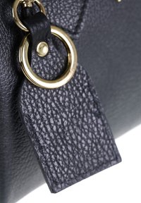 Angolo di una borsa a tracolla in pelle nera testurizzata con un anello in metallo dorato e un'etichetta in pelle. Presenta cuciture visibili e una superficie liscia.