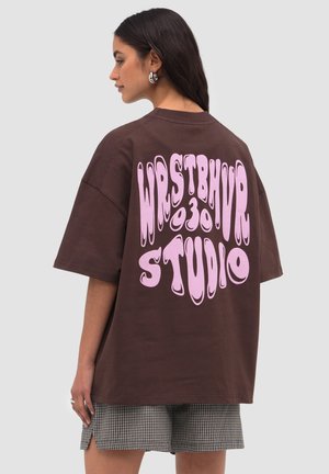 Mujer con cabello largo y oscuro, vistiendo una camiseta marrón oversize con texto psicodélico rosa "WRSTBHVR 30 STUDIO" en la espalda y pantalones cortos a cuadros.