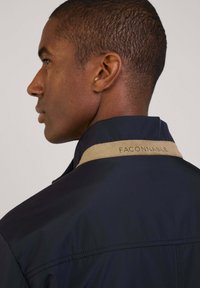 Chaqueta navy con cuello beige que presenta el bordado "FACONNABLE". Textura suave, corte entallado, costuras visibles en el hombro.