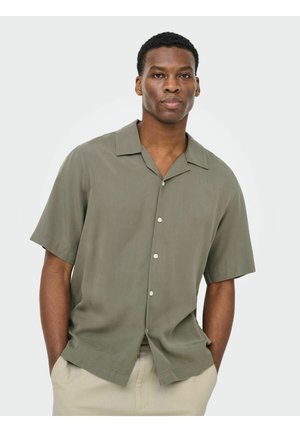 Homme portant une chemise ample à manches courtes olive à boutons et un pantalon beige, mains dans les poches, expression neutre, fond uni.
