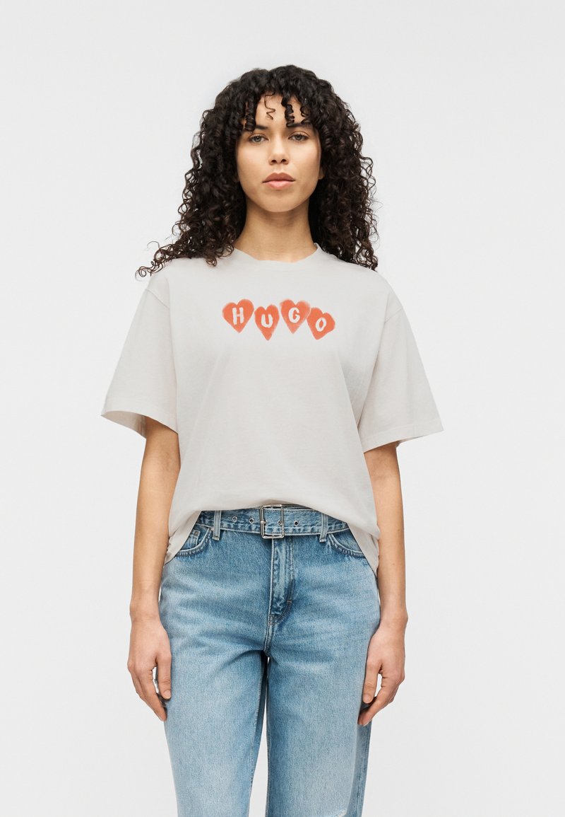 Ανοιχτό γκρι βαμβακερό μπλουζάκι με κόκκινες γραφικές καρδιές και το κείμενο "HUGO" στο κέντρο. Χαλαρή εφαρμογή με κοντά μανίκια.