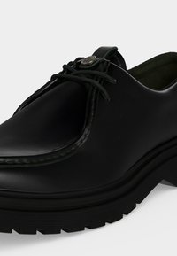 Chaussure habillée en cuir noir avec une semelle en caoutchouc épaisse, des lacets noirs et un petit bouton métallique rond près de la languette.