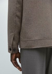 Brown wool-blend jacket with a straight cut, featuring a smooth texture and two black buttons at the cuff, paired with grey trousers.