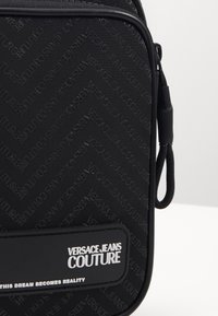 Svart väska med präglat "Versace Jeans Couture" logotypmönster, med en slät yta och en frontficka med silvertext och accenter.